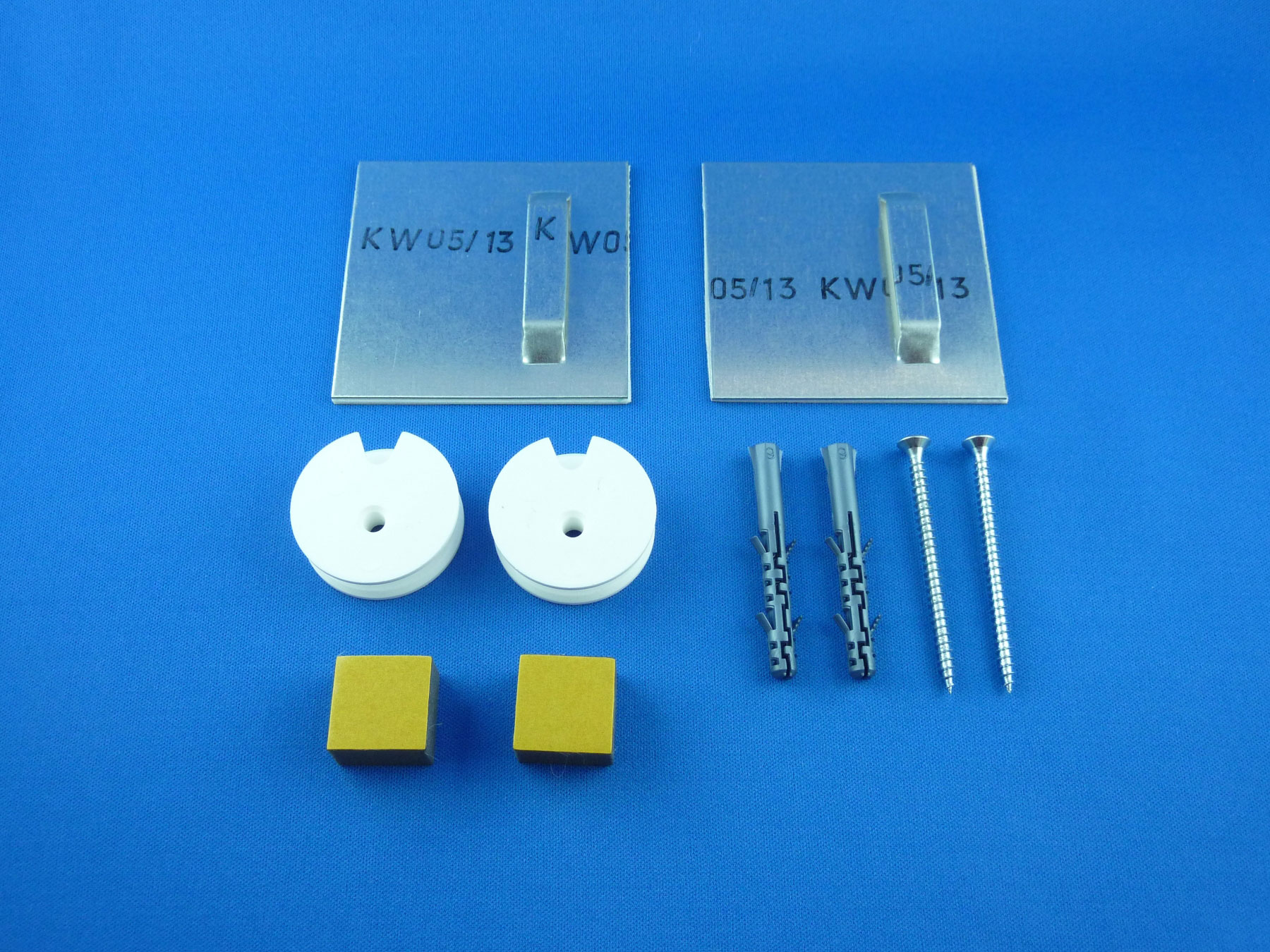 Ein Wandmontage-Set bestehend aus zwei Metallhalterungen, zwei Kunststoffrollen, zwei Dübeln, zwei Schrauben und zwei quadratischen Polstern auf blauem Hintergrund.