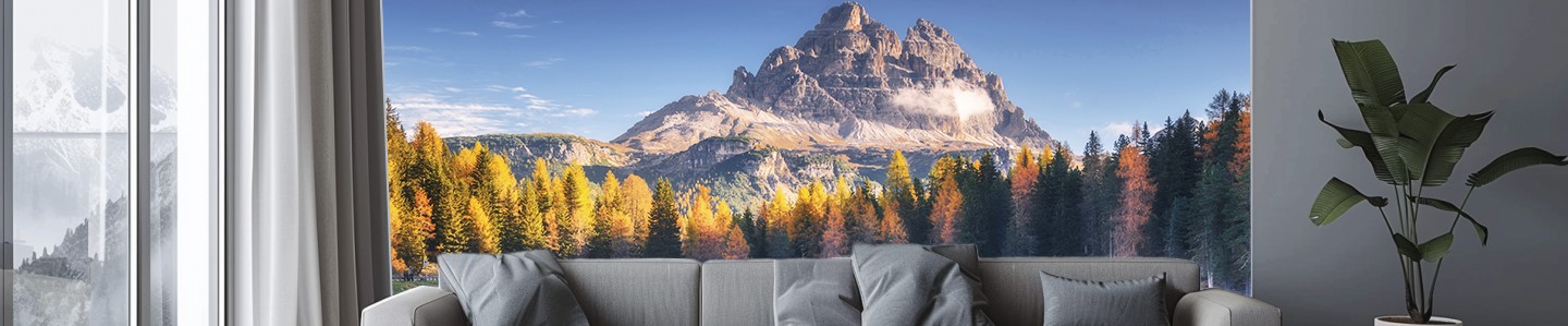 Wohnzimmer mit grauem Sofa, hellen Vorhängen und weitem Ausblick auf einen herbstlichen Wald und einen Berg im Hintergrund