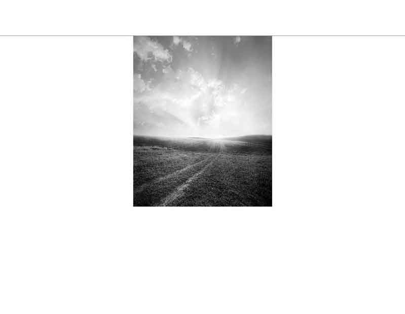 Ein schwarzweißes Bild zeigt eine weite Ebene unter einem wolkigen Himmel, durch den die Sonne strahlt.