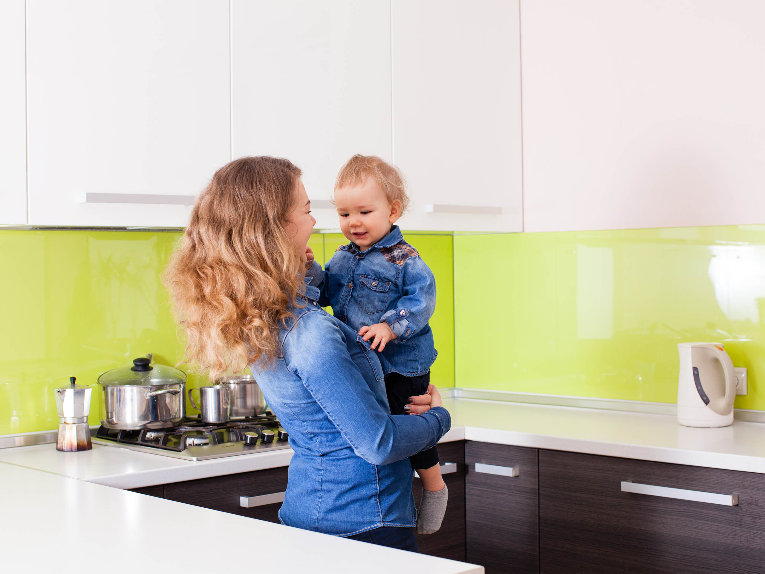Eine Frau mit lockigem Haar hält ihr kleines Kind in einer modernen Küche mit grüner Rückwand und weißem Wasserkocher.