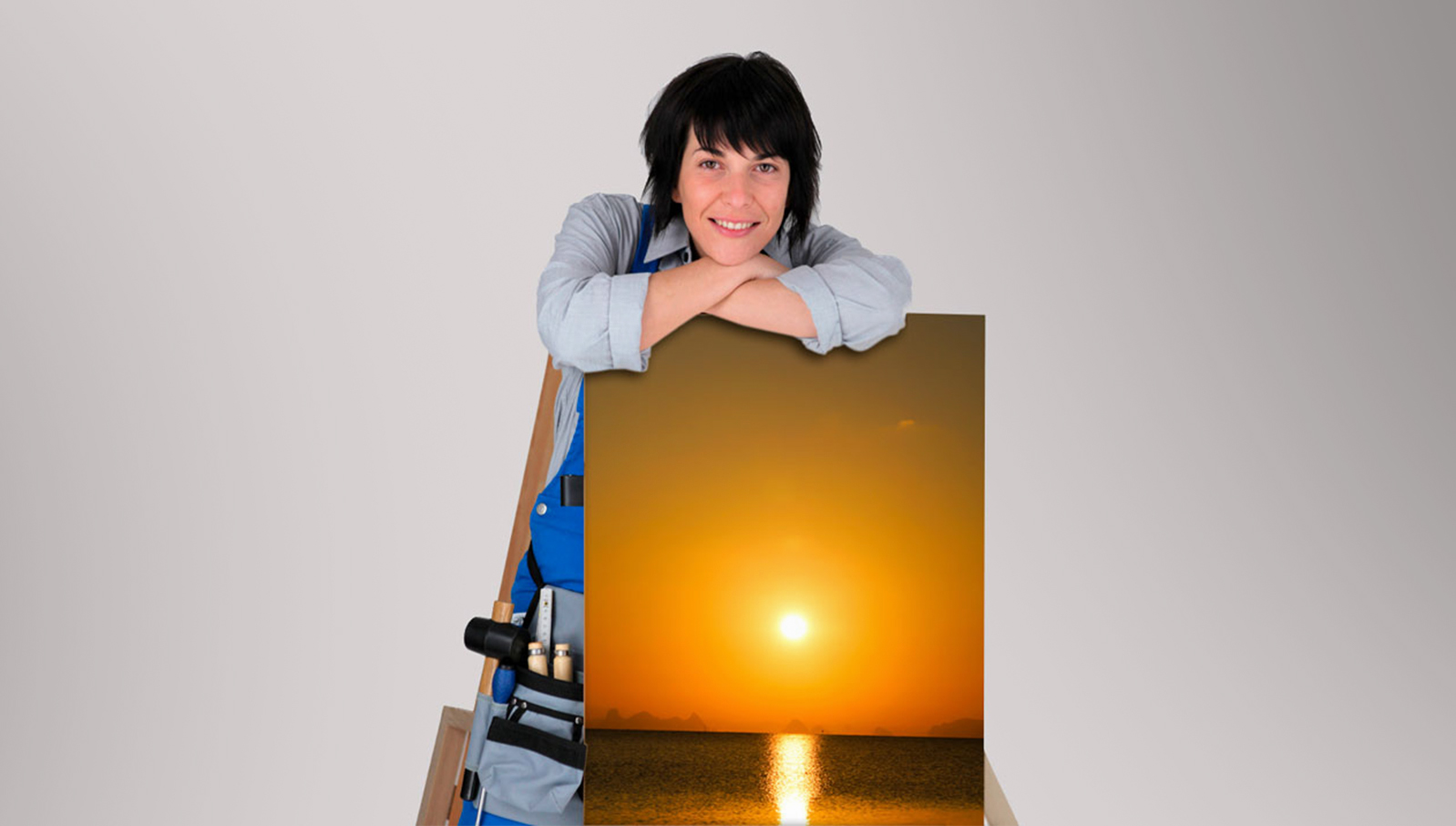 Eine Frau in Arbeitskleidung lehnt sich auf eine Leiter und hält ein Bild des Sonnenuntergangs über dem Meer, vor einem hellen Hintergrund.