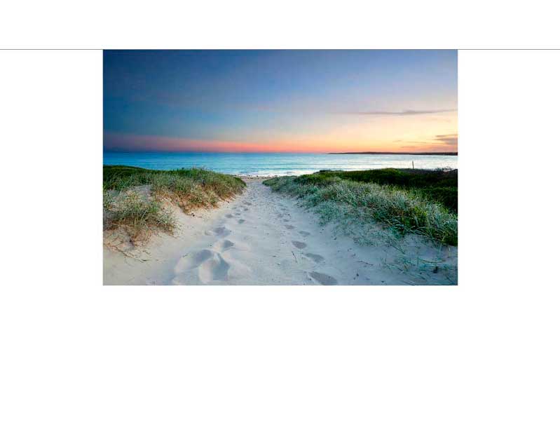 Ein malerischer Weg führt durch grasbewachsene Dünen zu einem ruhigen Strand bei Sonnenuntergang.