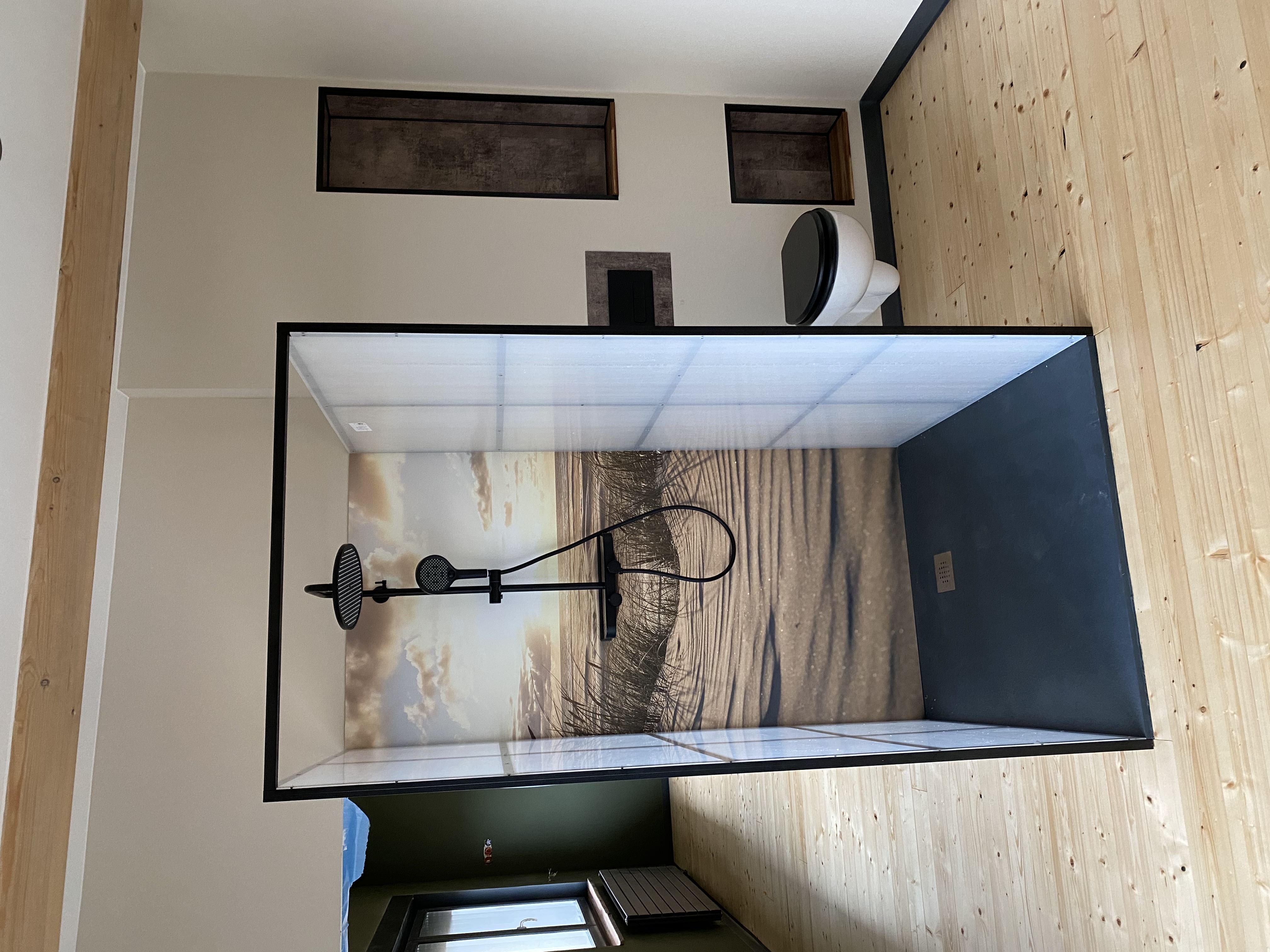 Ein Badezimmer mit einer modernen, freistehenden Dusche, deren Rückwand ein Motiv mit Strand und Meer zeigt, dazu ein hängendes WC auf Holzboden.
