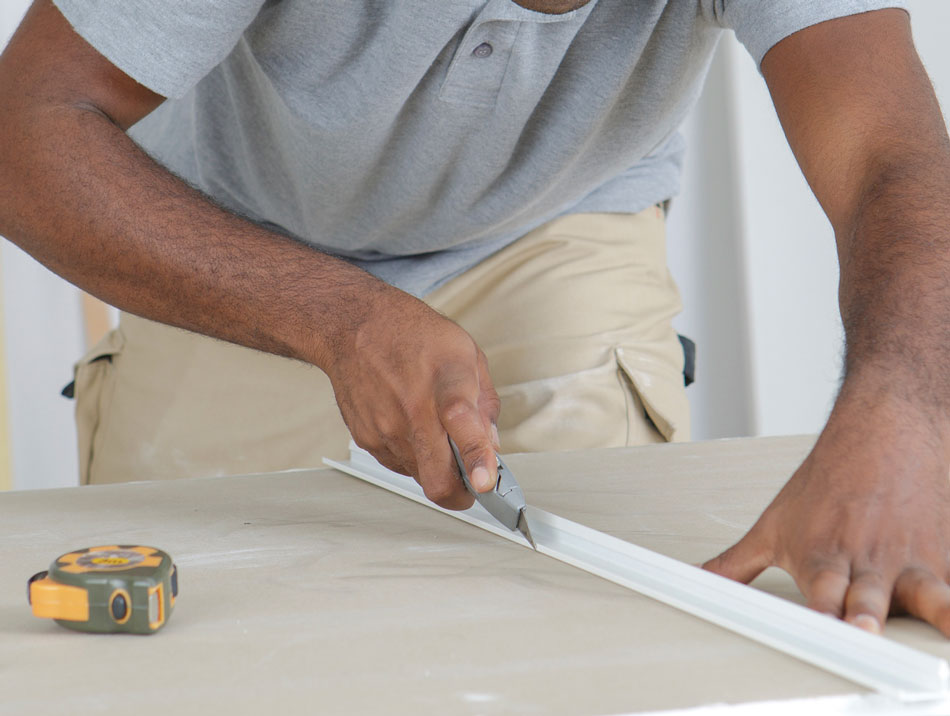 Nahaufnahme eines Mannes, der eine Plastikleiste mit einem Cutter-Messer zuschneidet. Auf dem Tisch liegt ein Maßband.